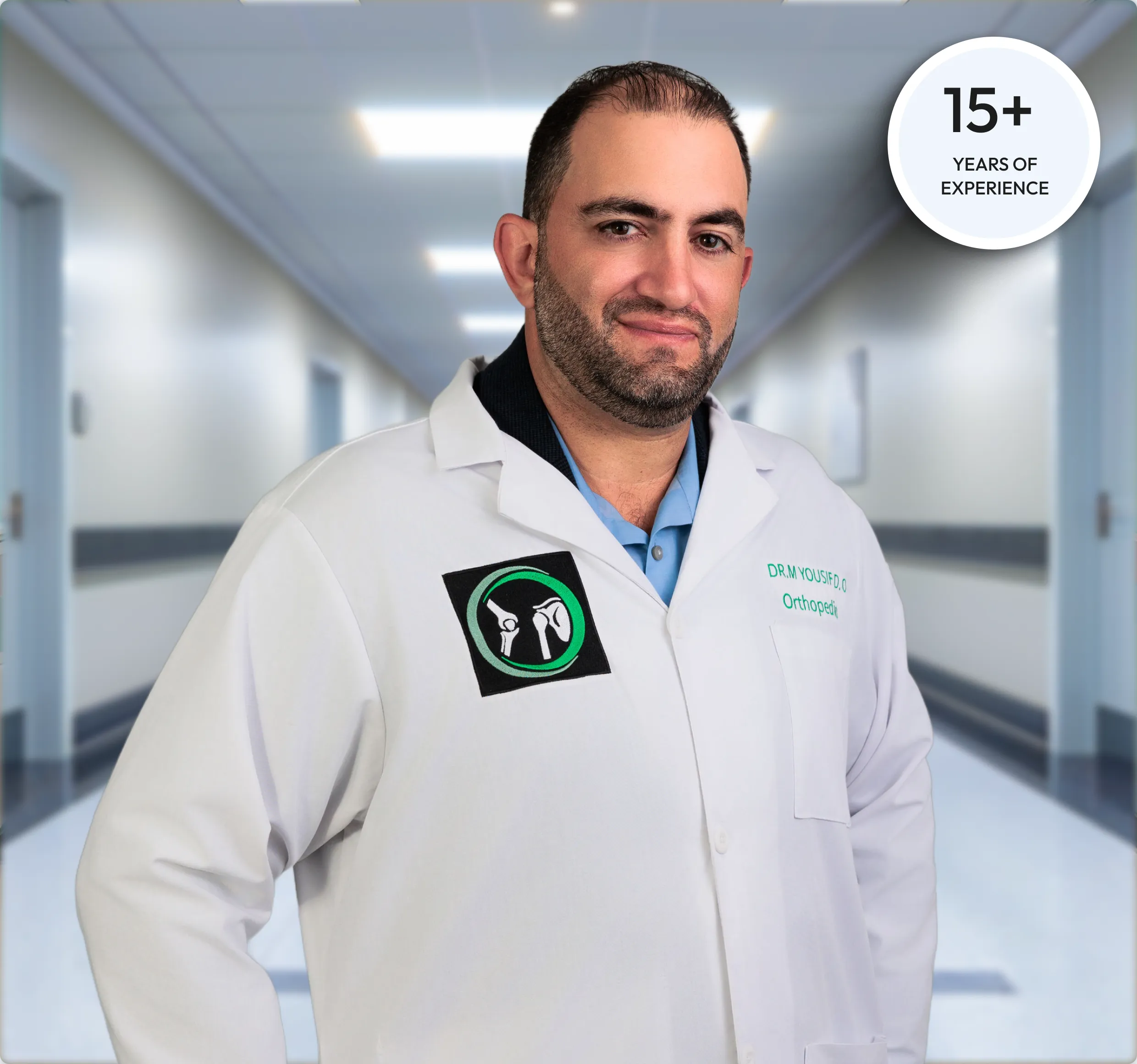 A male doctor in a white lab coat stands in the middle of a hospital hallway. The doctor, who has a beard and short dark hair, is smiling faintly and looking at the camera.