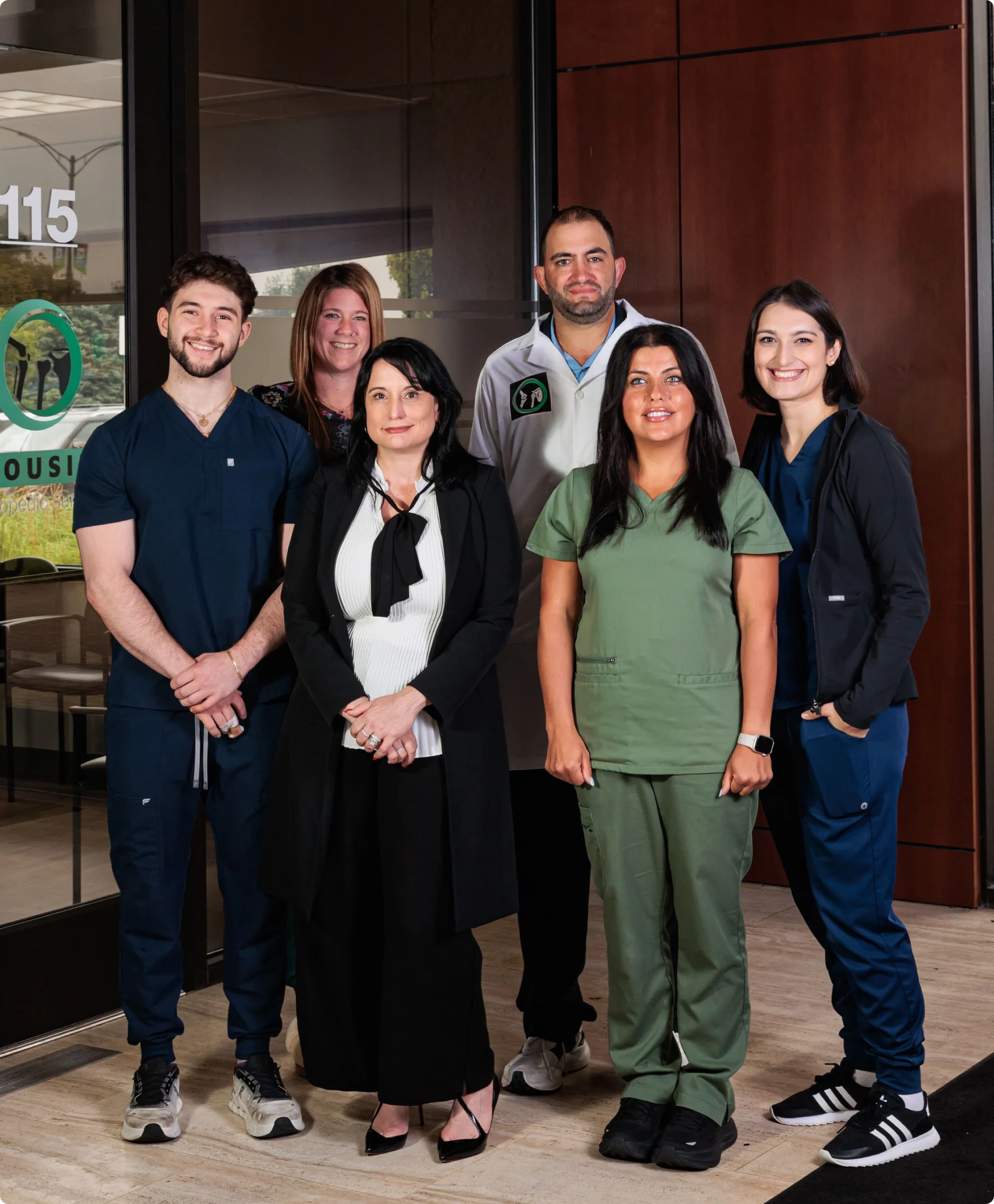 A professional group photo shows six people, four women and two men, standing and posing for the camera. The man on the far left and the woman on the far right are both in dark blue medical scrubs and smiling.