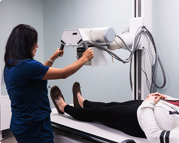 Medical technician operating an X-ray machine to take an image of a patient's lower body (legs/hips) in the orthopedic office.